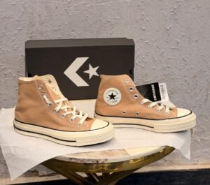 Beige high-top canvas-style sneakers with cream laces, metal eyelets, an off-white midsole with black pinstripes, and a dark brown patterned outsole