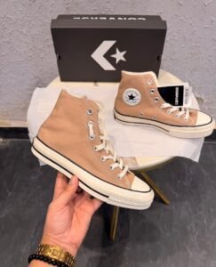 Beige high-top canvas-style sneakers with cream laces, metal eyelets, an off-white midsole with black pinstripes, and a dark brown patterned outsole