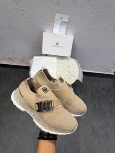 Beige slip-on sneakers with a metallic silver-tone buckle, broad front strap, and sculpted white outsole