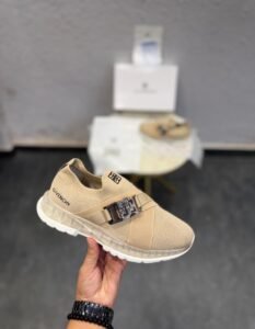 Beige slip-on sneakers with a metallic silver-tone buckle, broad front strap, and sculpted white outsole