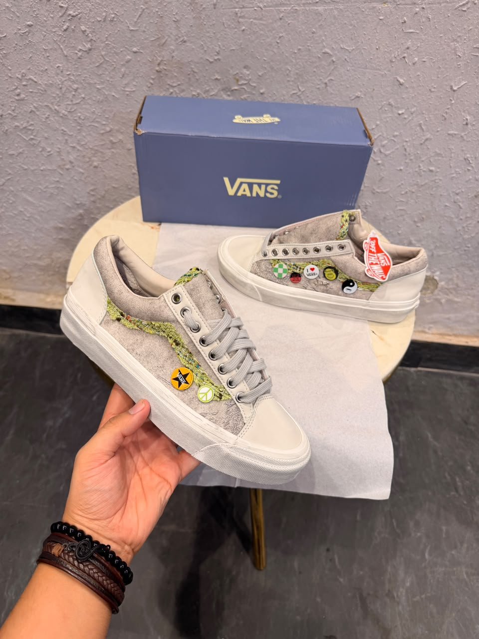 Beige low-top sneakers with green textured side detailing, decorative circular badges, cream toe panels, and a black patterned outsole