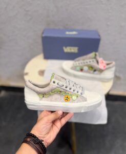 Beige low-top sneakers with green textured side detailing, decorative circular badges, cream toe panels, and a black patterned outsole