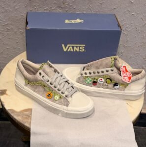 Beige low-top sneakers with green textured side detailing, decorative circular badges, cream toe panels, and a black patterned outsole