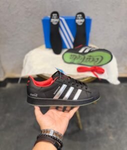 Black retro low-top sneakers with silver-toned stripes, red accents, a textured shell-shaped toe, and a green-toned outsole