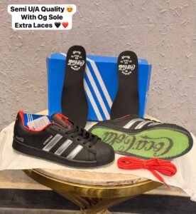Black retro low-top sneakers with silver-toned stripes, red accents, a textured shell-shaped toe, and a green-toned outsole