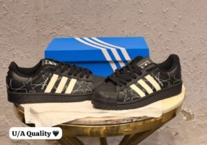 Black low-top sneakers with cream-coloured stripes, decorative star stitching, and a textured shell-shaped toe cap